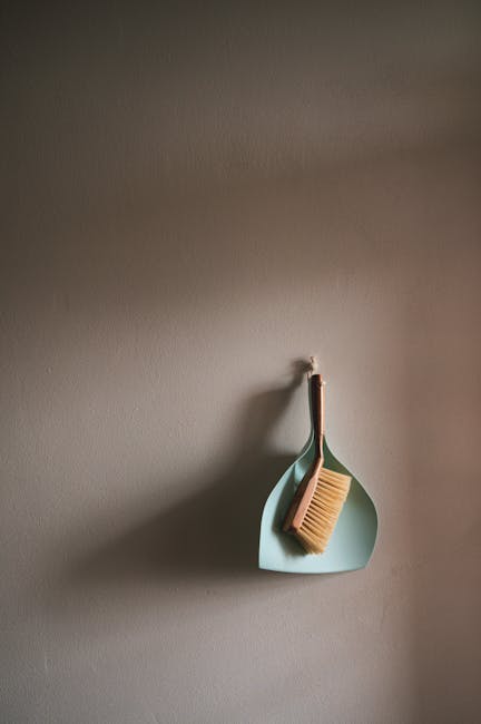 A cleaning brush with a wooden handle and natural bristles hangs on a small hook attached to a plain, beige wall. Beneath the brush, a pale blue silicone dish or holder is mounted, providing a space f