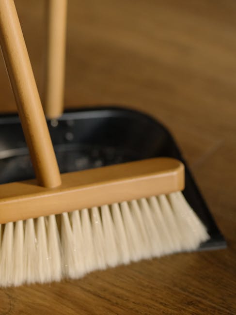 A cleaning brush with a wooden handle and natural bristles hangs on a small hook attached to a plain, beige wall. Beneath the brush, a pale blue silicone dish or holder is mounted, providing a space f